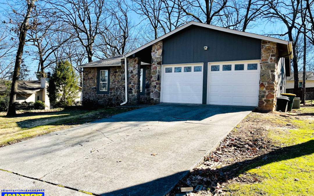 Handsome refurbished home in West Little Rock, featuring a stone facade, two-car garage, and a sloped driveway surrounded by trees.