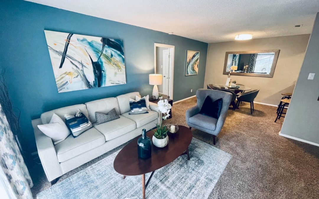 Modern living room and dining area in NLR Hilltop Community, featuring a light gray sofa with decorative pillows, a round coffee table with plants, and a stylish gray armchair, complemented by blue accent walls and artwork.