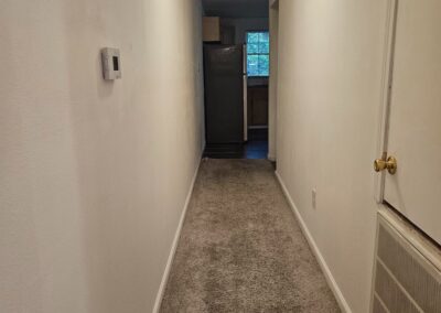 Hallway of a newly renovated duplex in Midtown Little Rock, featuring beige walls, carpeted flooring, and a view of a refrigerator and kitchen area at the end.