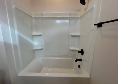 Modern bathroom tub and shower combo with white acrylic surround, black fixtures, and built-in shelves, showcasing features of a brand-new townhome in the Argenta District.
