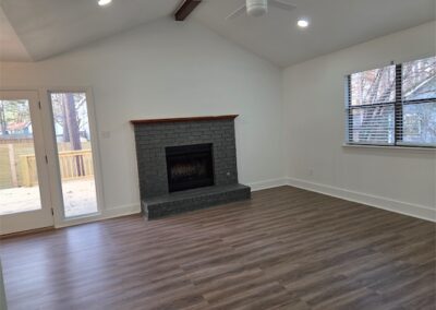 Spacious living room featuring new hardwood flooring, modern ceiling fan, stone fireplace, large windows, and access to a private fenced backyard.
