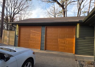 Attached two-car garage with modern orange doors and contemporary lighting, adjacent to a driveway and surrounded by trees, showcasing the home's exterior in West Little Rock.